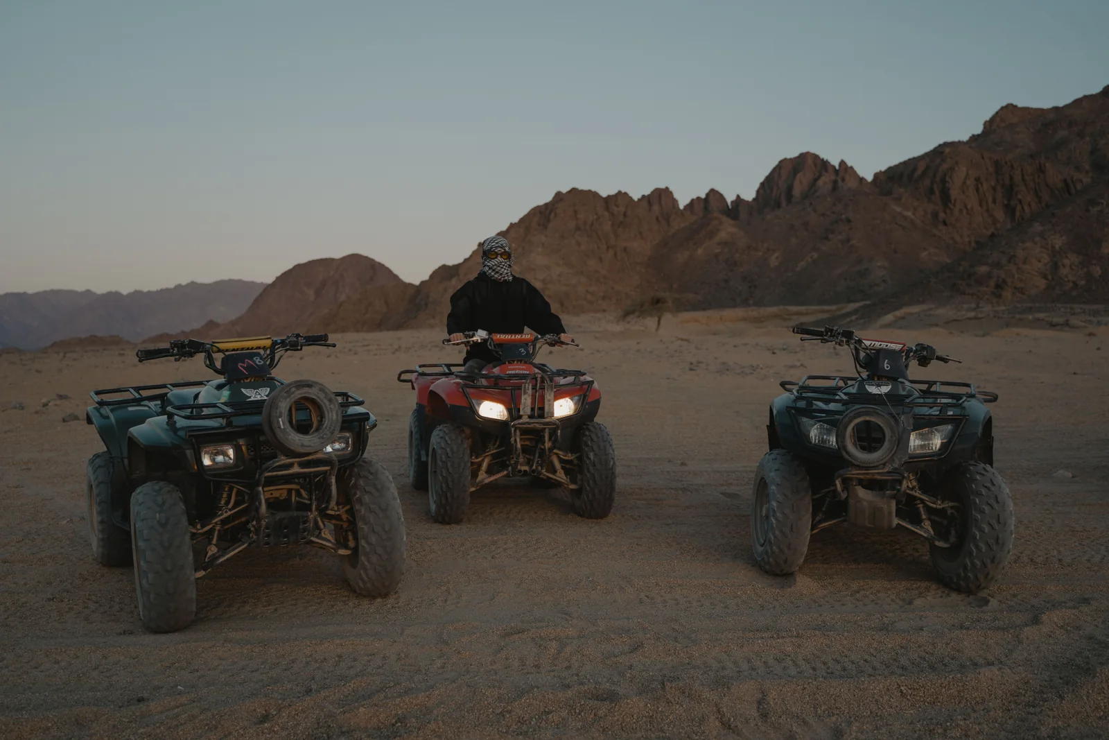 Three quad bikes and a rider on rocky desert terrain with mountains behind
