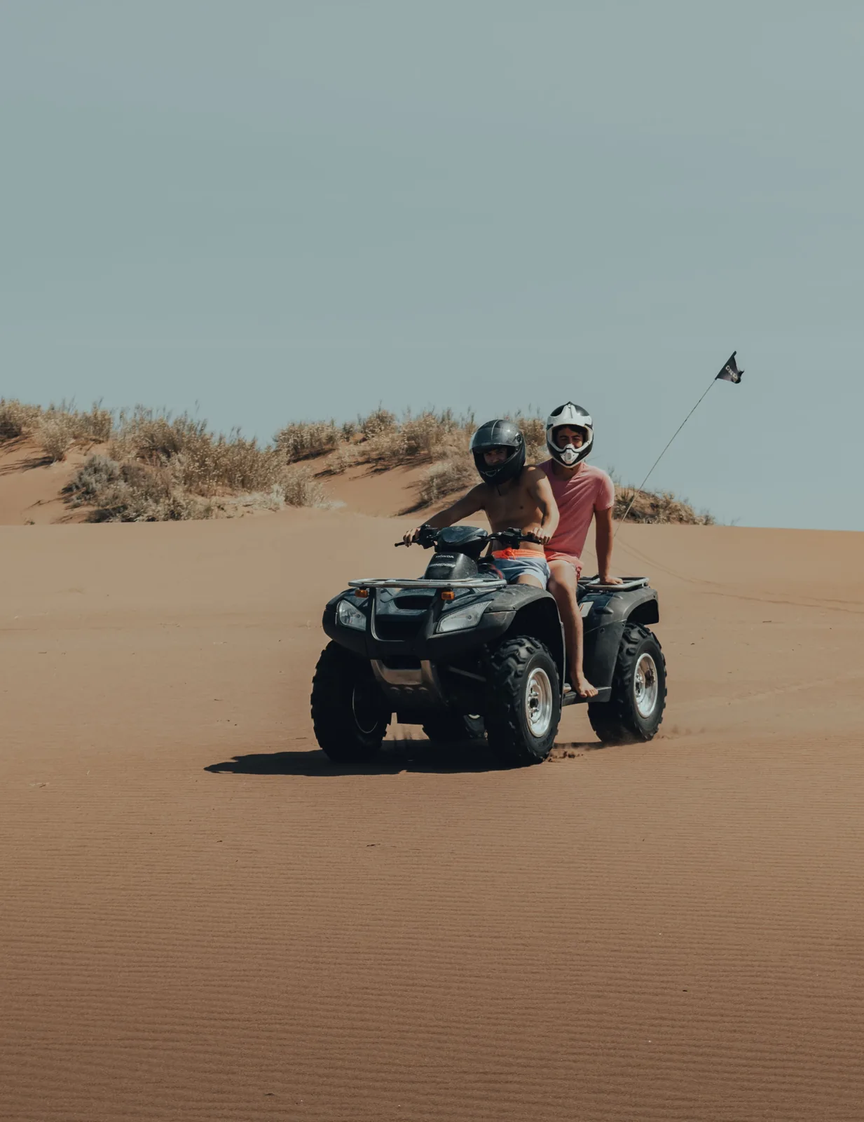 Two helmeted riders on a quad bike crossing sandy dunes